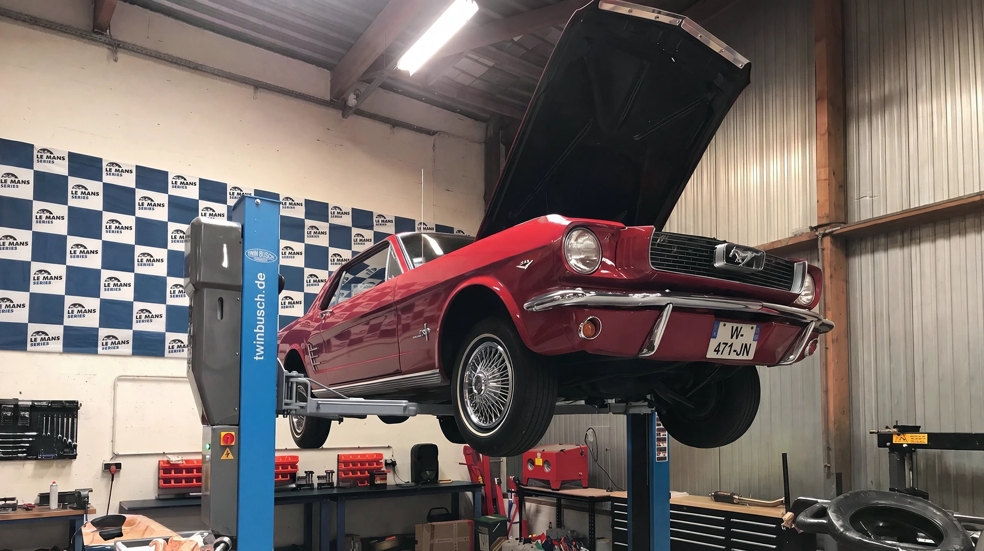 Atelier automobile intérieur spécialisé en restauration de véhicules de collection. Au centre, une voiture classique vintage (années 1960-1970) de prestige ou de collection est stationnée sur un pont élévateur hydraulique ou au sol, partiellement démontée : capot ouvert, portes retirées ou ouvertes, révélant le châssis et les composants mécaniques en cours de restauration. En arrière-plan visible : établis avec outillage spécialisé de restauration, pièces détachées d'époque rangées sur étagères, équipement de diagnostic, systèmes d'aspiration de poussière, bacs de travail. Ambiance d'atelier méticuleux éclairé aux LED, sol époxy, soulignant l'expertise technique et le caractère précis de la restauration de collection. Aucun personnage visible. Photo de qualité professionnelle.